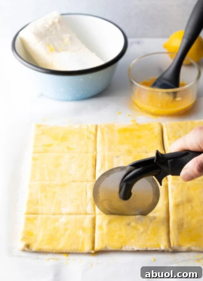 Hand using a pizza cutter to precisely slice a sheet of puff pastry into uniform rectangles.
