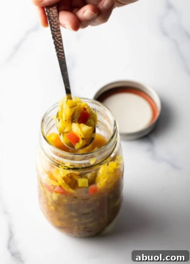 Top down view glass jar of chow chow. The lid is off and to the right side, a metal spoon is in the jar showing a scoop to camera.