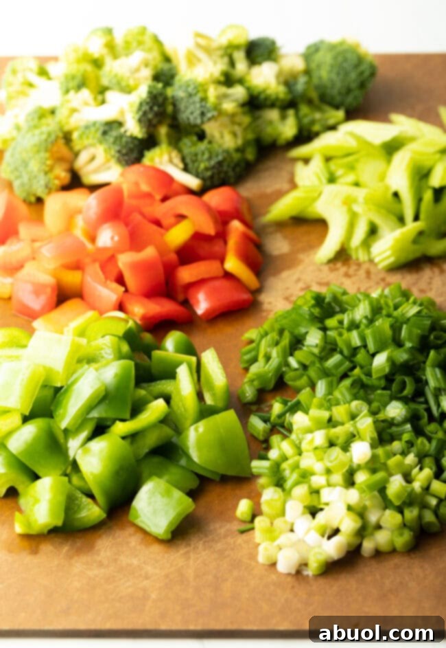 A cutting board neatly arranged with piles of prepped green bell pepper, red bell pepper, scallions, celery, and broccoli florets, ready for cooking.