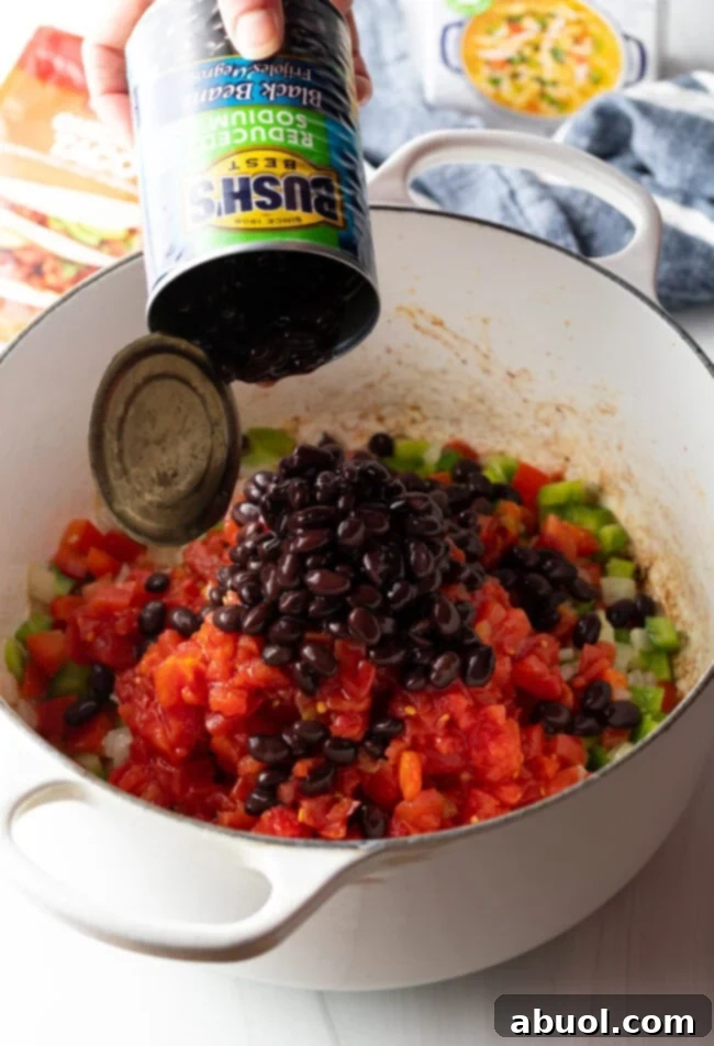 A hand adding canned black beans to a pot with sautéed vegetables, corn, and broth.
