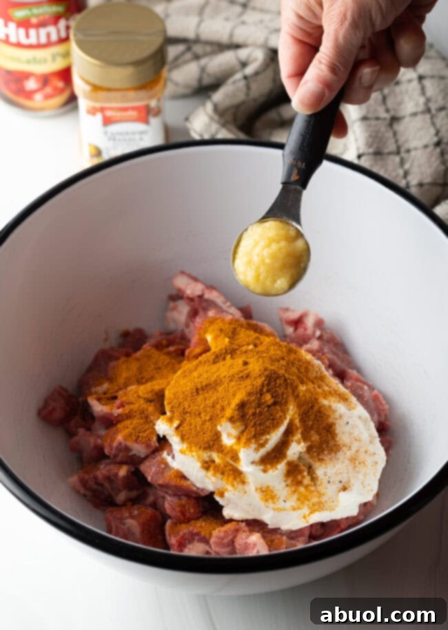 Creamy Lamb Tikka Masala 6 Hand with measuring spoon adding spices to yogurt and pieces of lamb meat in a white bowl.
