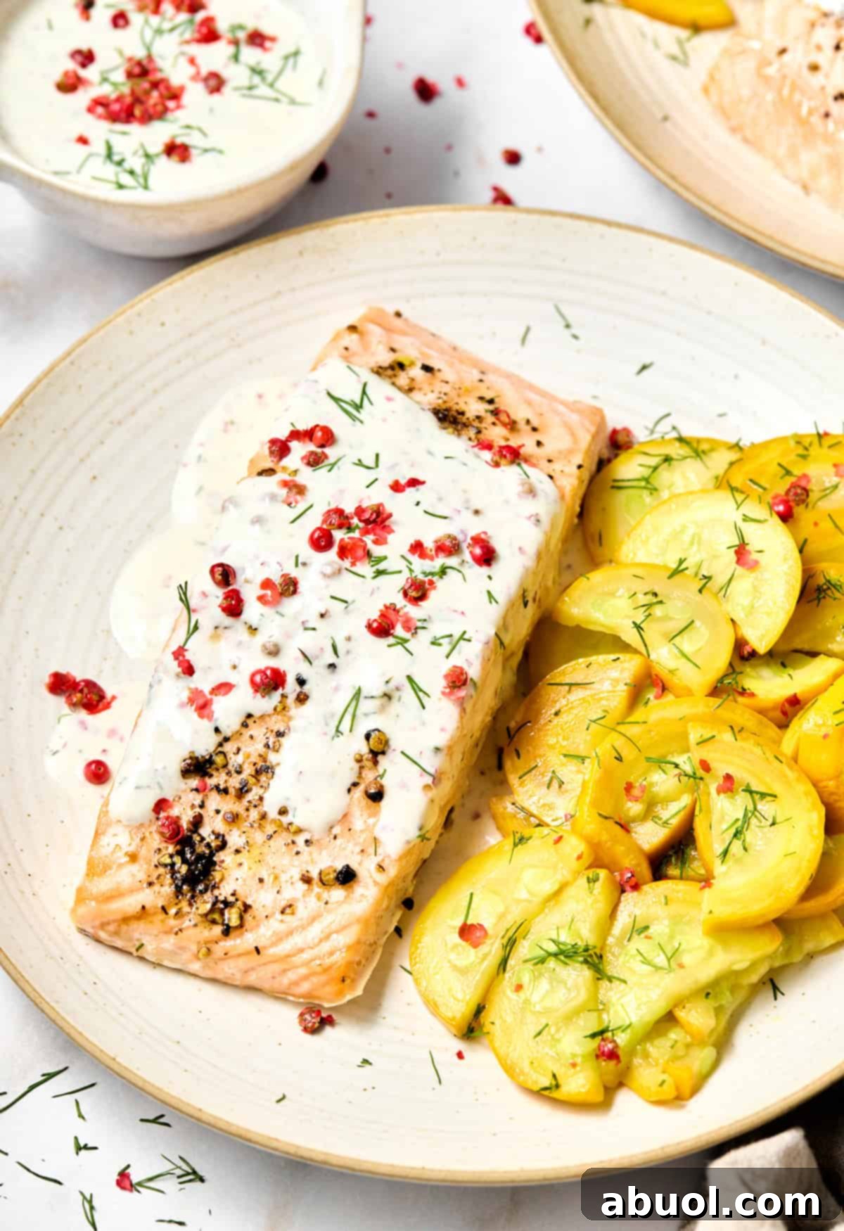 Baked salmon on a plate with pink peppercorn sauce and fresh whole pink peppercorns on top. A serving of yellow cooked squash is on the side.