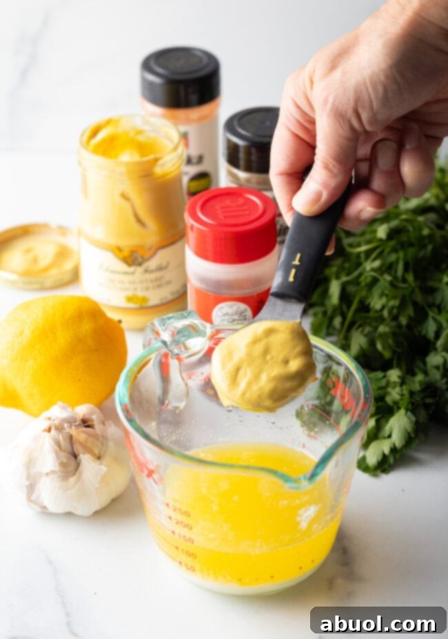 Hand with metal measuring spoon of Dijon mustard, adding to the melted butter in a glass measuring pitcher.