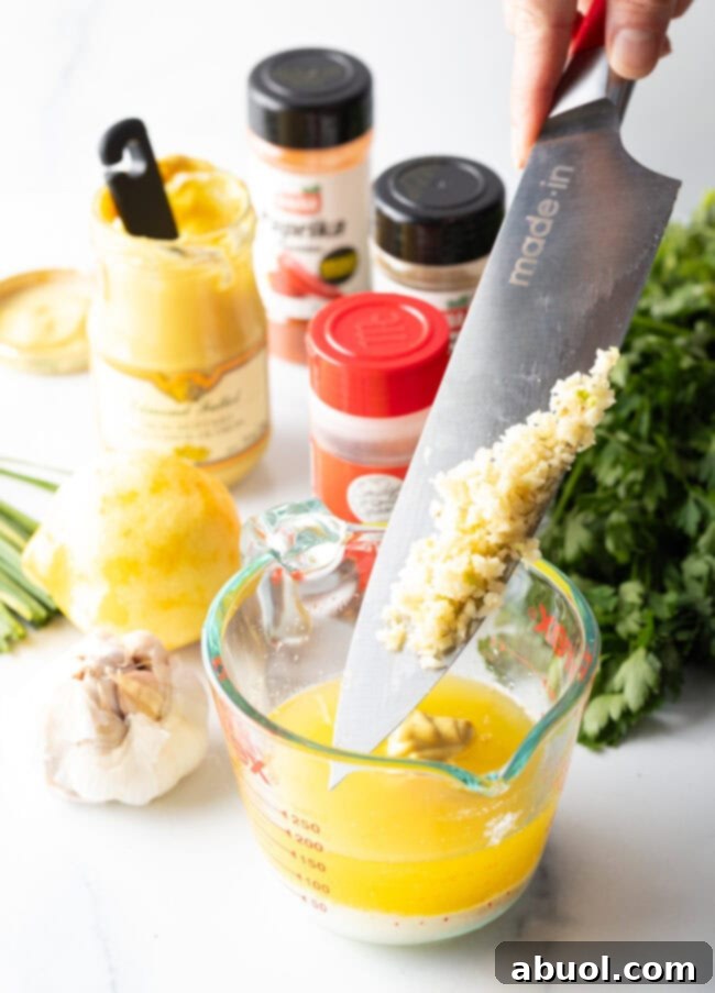Knife with minced garlic, adding garlic to melted butter in a glass measuring pitcher.