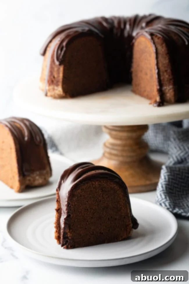 Chocolate Glaze for Pound Cake - Thick slice of chocolate sour cream pound cake with drizzle of glaze on a white plate. The full cake is on a white cake stand in the background.