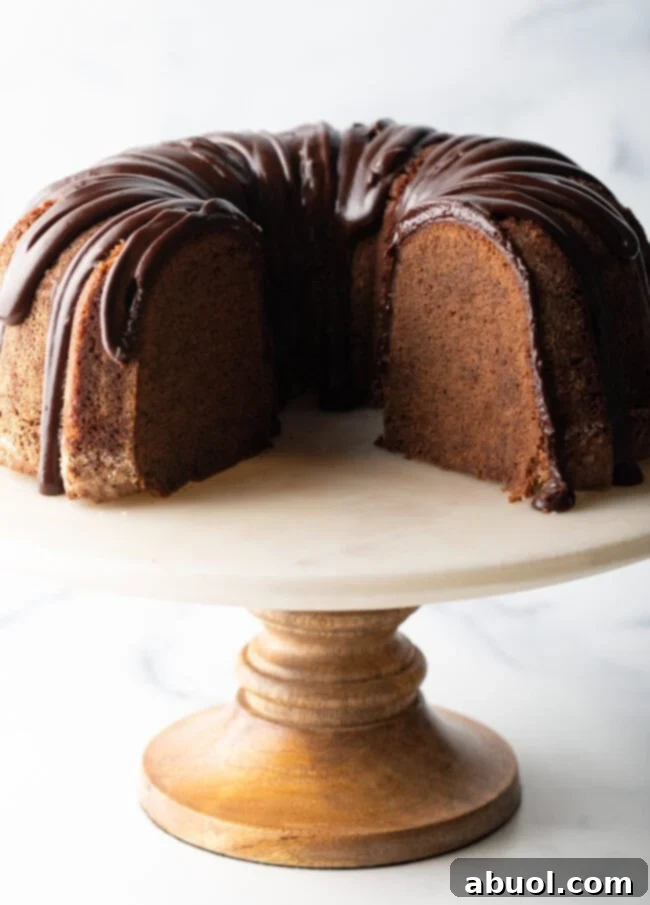 Chocolate sour cream bundt cake with a thick chocolate glaze drizzled on top. The cake is on a white stand and has a portion removed to show the tender inside crumb.