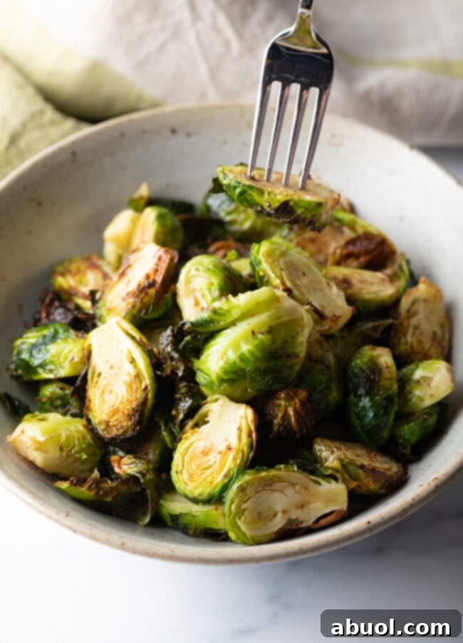 Fork holding a brussel sprout half over a white bowl of air fried sprouts.