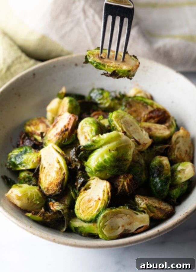 Fork holding a brussel sprout half over a white bowl of air fried sprouts.
