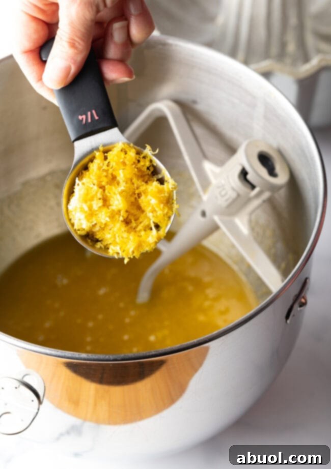 Golden Lemon Olive Oil Cake 6 A hand holding a metal measuring cup, zesting a fresh lemon directly over a mixing bowl, capturing the essence.