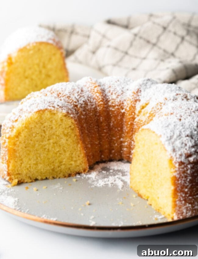 Golden Lemon Olive Oil Cake 10 A lemon olive oil bundt cake, dusted with powdered sugar, on a white plate. A large slice has been removed, revealing its inviting yellow, moist crumb.