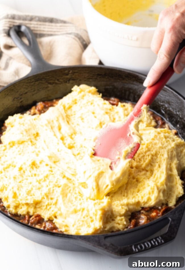 A hand using a spatula to spread the yellow cake batter evenly over the pecan mixture in a cast iron skillet, ready for baking.