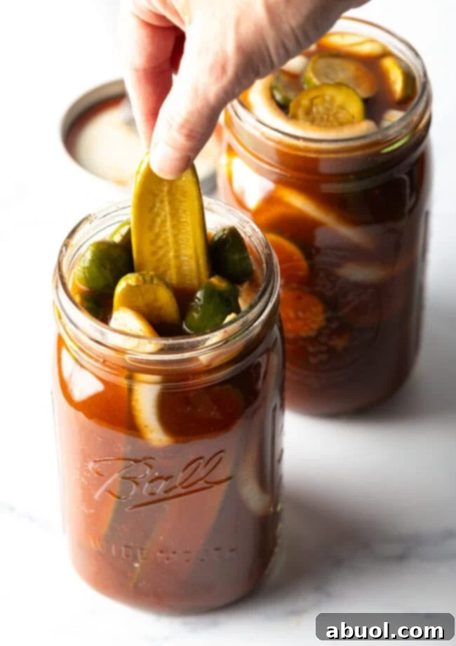 Two large mason jars of chamoy pickles. The tops are off of the jars, and a long pickle is being pulled out of one jar.