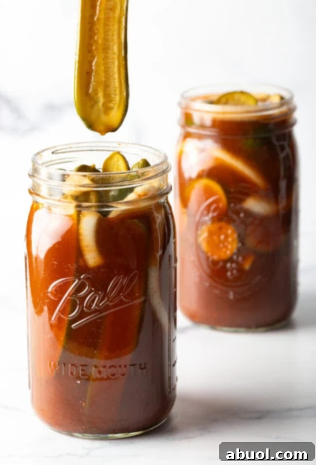 Two large mason jars of chamoy pickles. The tops are off of the jars, and a long pickle is being pulled out of one jar.