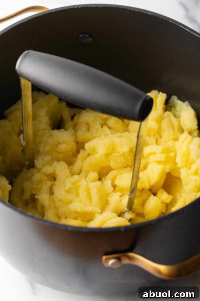 A hand masher is diligently used to thoroughly mash cooked yellow potatoes in a large pot.