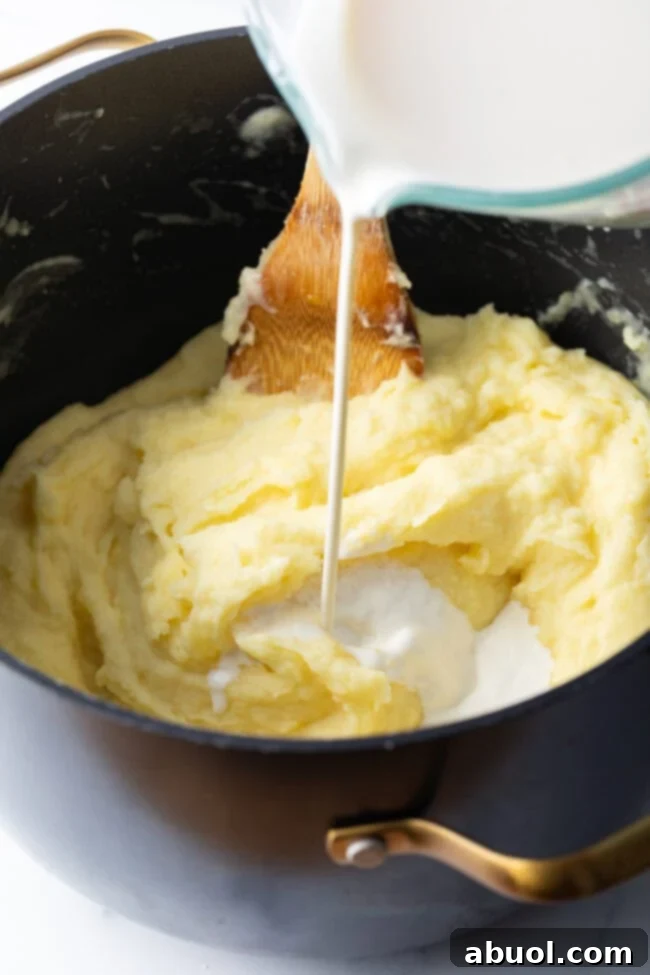 Heavy cream is being poured into a large pot filled with cooked, mashed potatoes, ready for stirring.