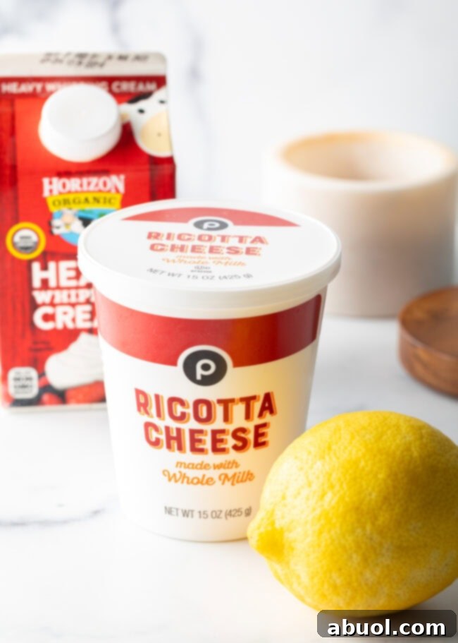 The simple yet essential ingredients for whipped ricotta: a tub of whole milk ricotta cheese, a carton of heavy cream, and a vibrant whole lemon on a white background, ready for preparation.