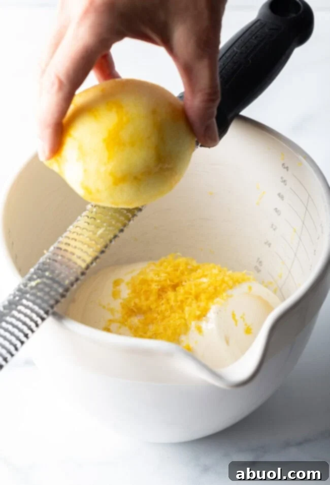 A hand using a lemon zester to grate fresh lemon zest directly into a white mixing bowl, where ricotta cheese and cream are already waiting, illustrating the first step of the recipe.