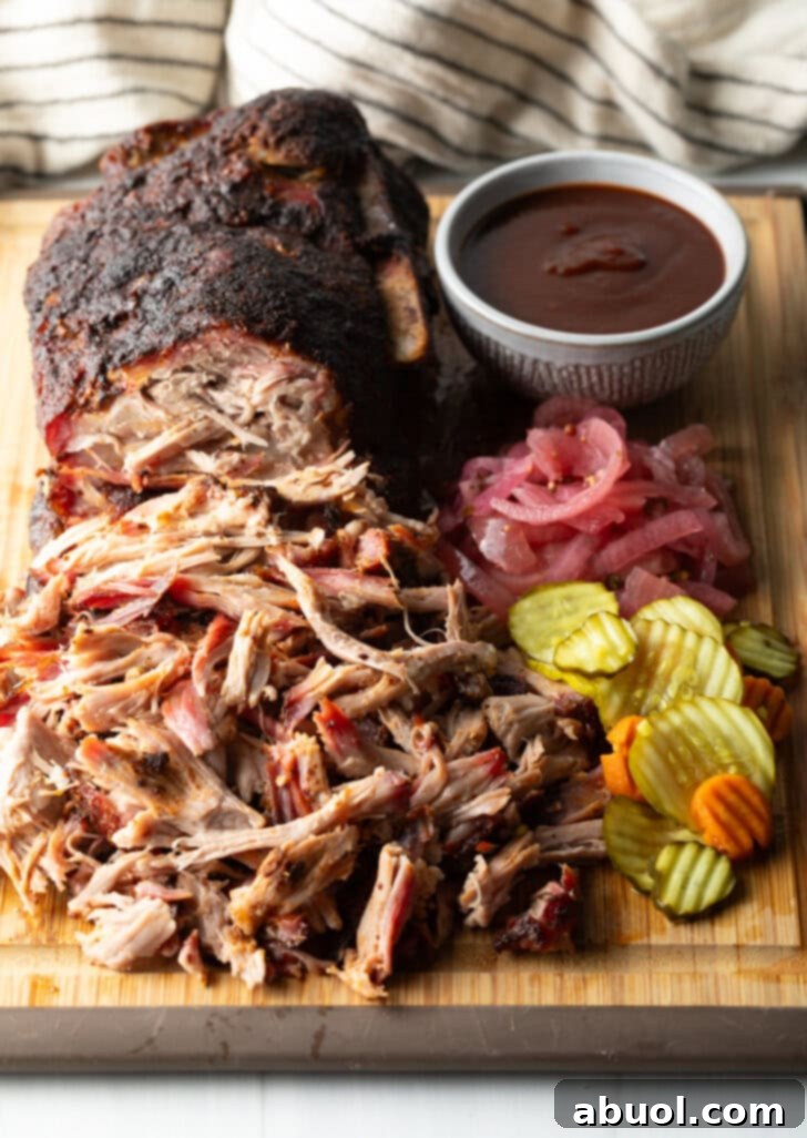 Shredded smoked pork butt on a cutting board, with ramekin of bbq sauce, stack of pickles, and pickled red onions.