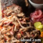 Shredded smoked pork butt on a cutting board, with ramekin of bbq sauce, stack of pickles, and pickled red onions.