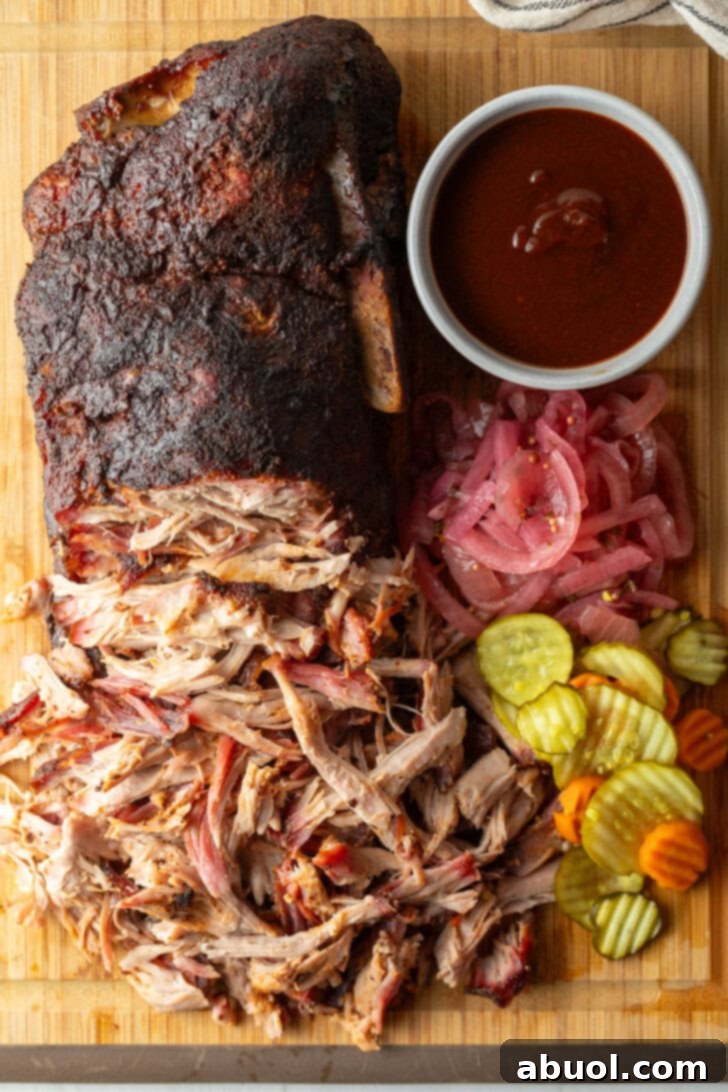 Shredded smoked pork butt on a cutting board, with ramekin of bbq sauce, stack of pickles, and pickled red onions.