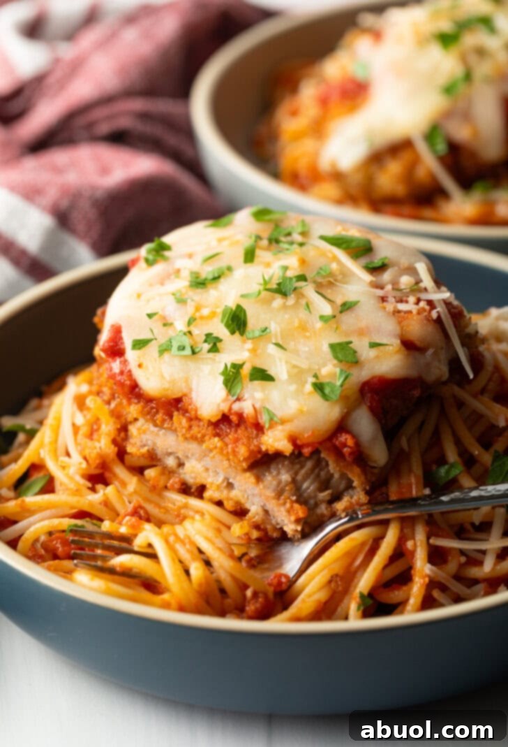 A cutlet of veal parmesan on top of spaghetti. A bite has been removed from the veal to show the juicy inside.