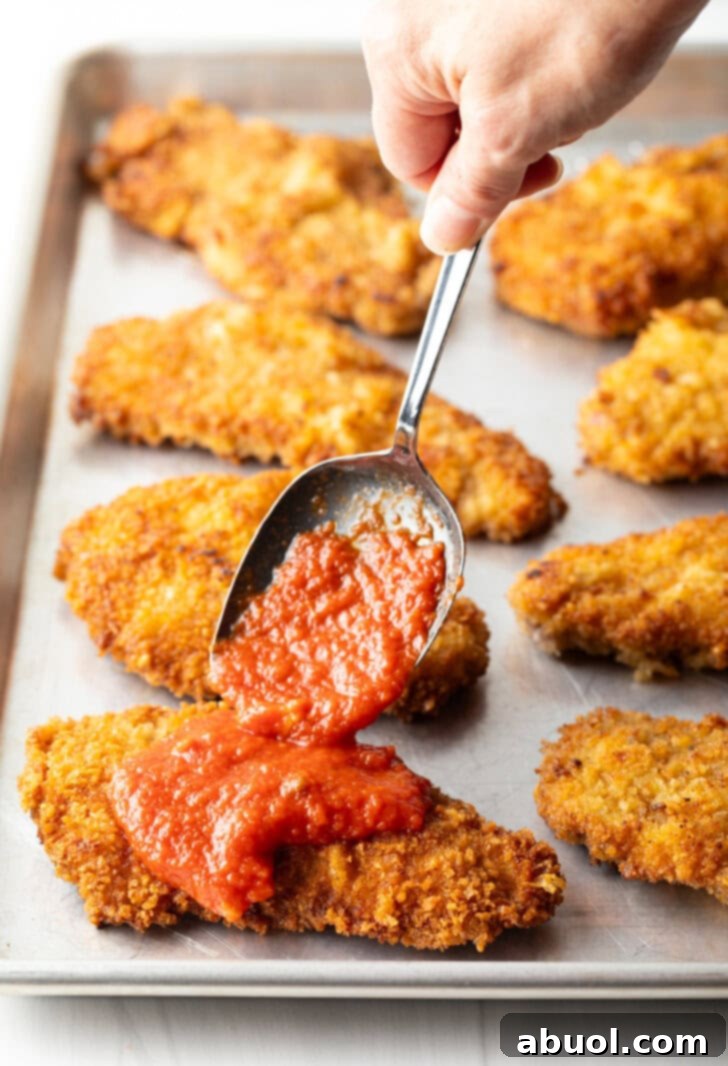 Hand with metal spoon adding marinara sauce on top of a breaded veal cutlet.