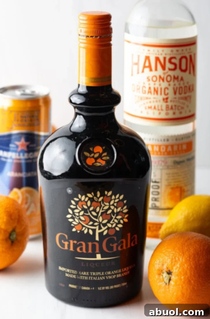 Ultimate Orange Crush Cocktail 5 Recipe ingredients: Bottle of Grangala in the front, fresh oranges, bottle of vodka, and can of orange soda in the background.