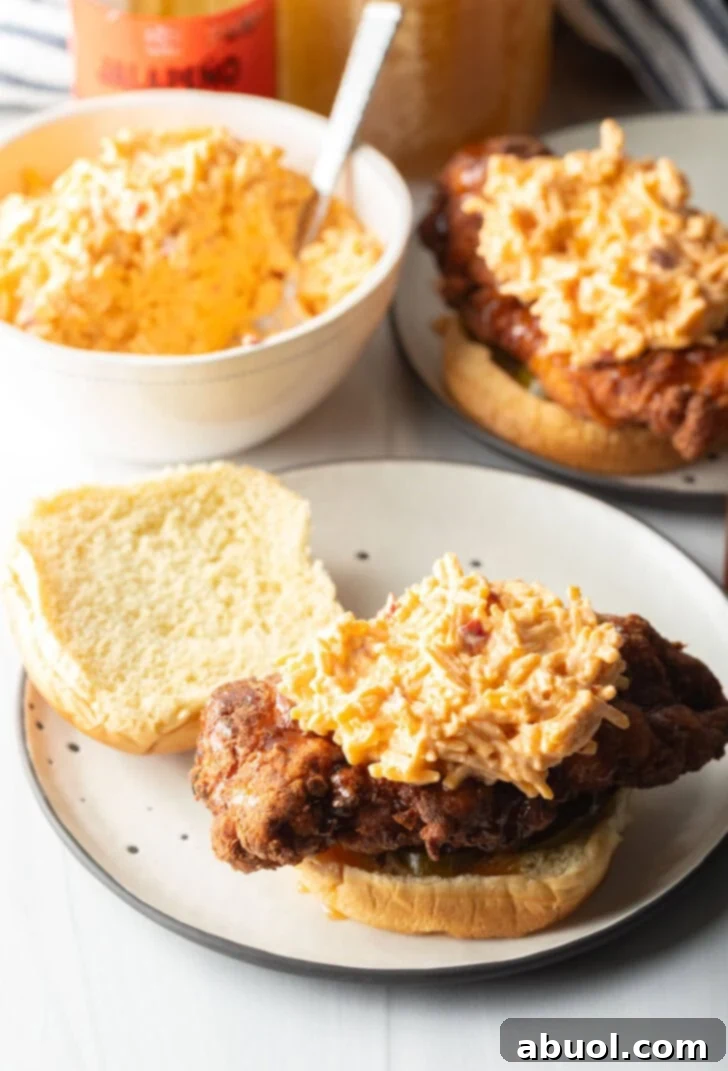 An open-faced fried chicken breast sandwich showcasing a generous serving of rich, creamy pimento cheese spread on the golden chicken, layered over pickled jalapeños on a bun.