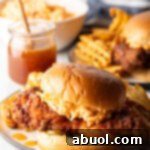 Honey pepper pimento chicken sandwich on a plate. In the background is a jar of honey, and second sandwich with waffle fries.