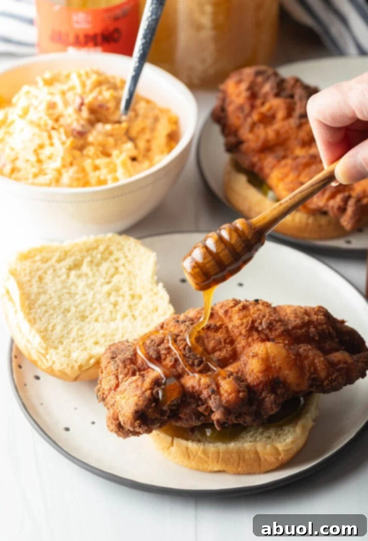 A stream of golden honey gracefully drizzles over a crispy breaded chicken breast, already placed on the bottom half of a burger bun with jalapeños, adding a sweet and spicy touch.