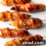 6 skewered bacon wrapped chicken tenders coated with hot sauce on a white plate.