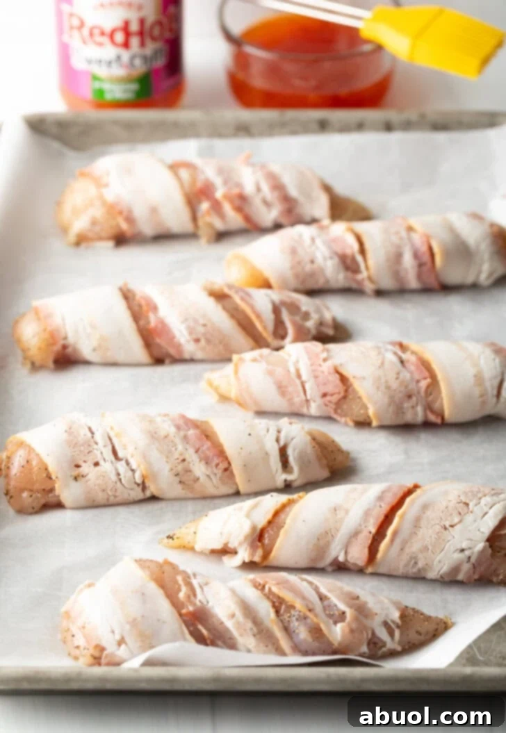 Mouthwatering Bacon-Wrapped Chicken 6 6 raw chicken breast tenders wrapped in raw bacon, placed on a baking sheet with white parchment paper.