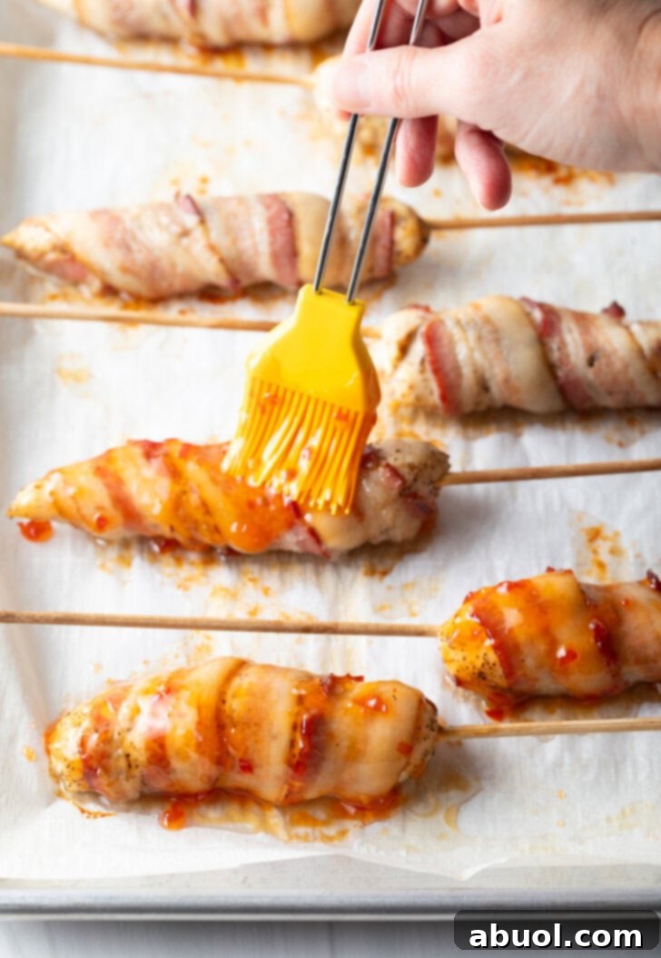Mouthwatering Bacon-Wrapped Chicken 7 6 raw chicken tender wrapped in bacon on a baking sheet with parchment paper. A yellow basting brush is brushing on hot sauce.