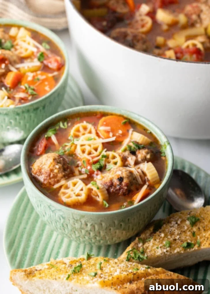 A vibrant green bowl filled with Italian meatball soup, showcasing wheel-shaped pasta, thinly sliced carrots, and perfectly browned meatballs in a rich, deep red tomato broth.