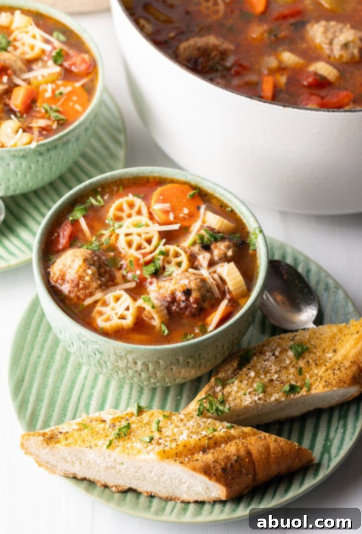 A vibrant green bowl of Italian meatball soup, generously laden with wagon wheel pasta, bright carrot slices, and succulent meatballs in a rich tomato broth. Two perfectly toasted baguette slices rest on the green plate beneath the soup bowl, ready for dipping.