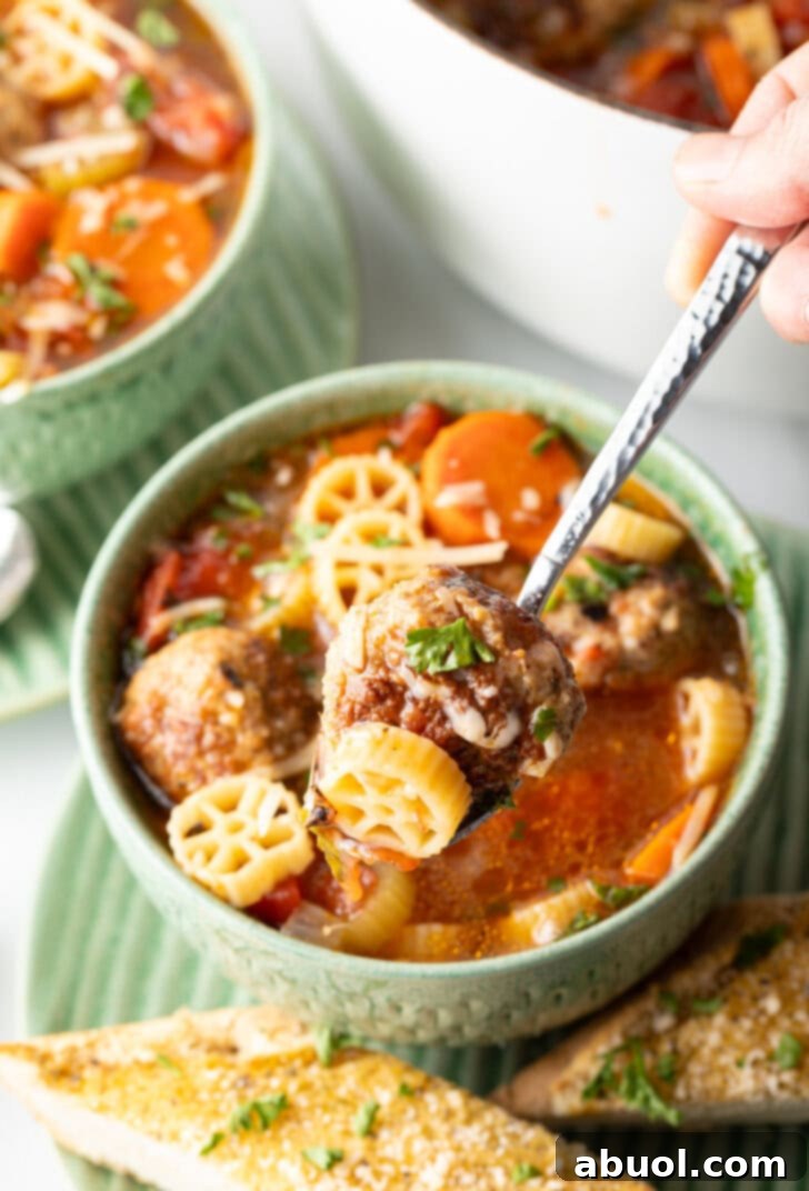 A vibrant green bowl of Italian meatball soup, brimming with wheel-shaped pasta, sliced carrots, and tender meatballs. A metal spoon gracefully lifts a bite of the soup towards the camera, highlighting the hearty ingredients.