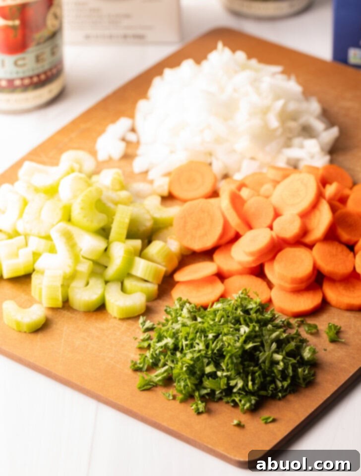 A wooden cutting board displaying neatly arranged piles of ingredients: thinly sliced carrots, diced celery, chopped onion, and fresh parsley, all ready for the soup.