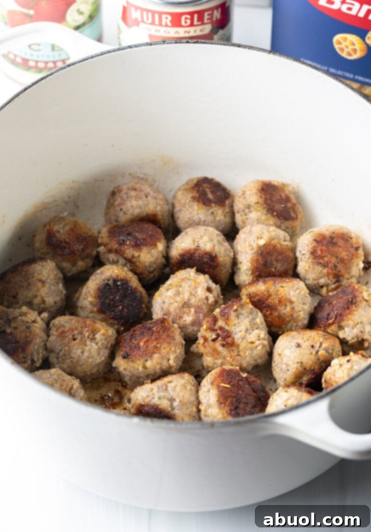 Perfectly seared and lightly browned meatballs rest in the bottom of a large white soup pot, having developed a flavorful crust.