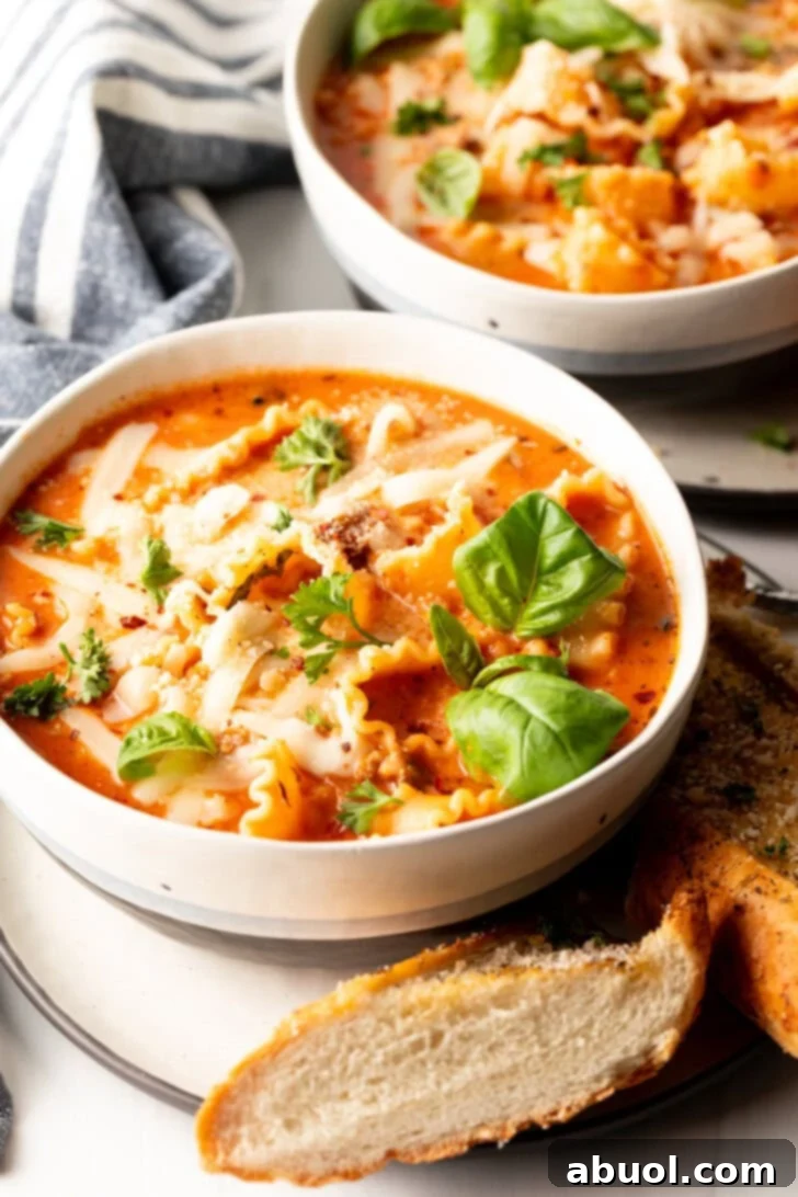 No-Fuss Lasagna Soup 2 White bowl loaded with lasagna soup, garnished with two fresh basil leaves. A slice of baguette is on a white plate under the bowl.