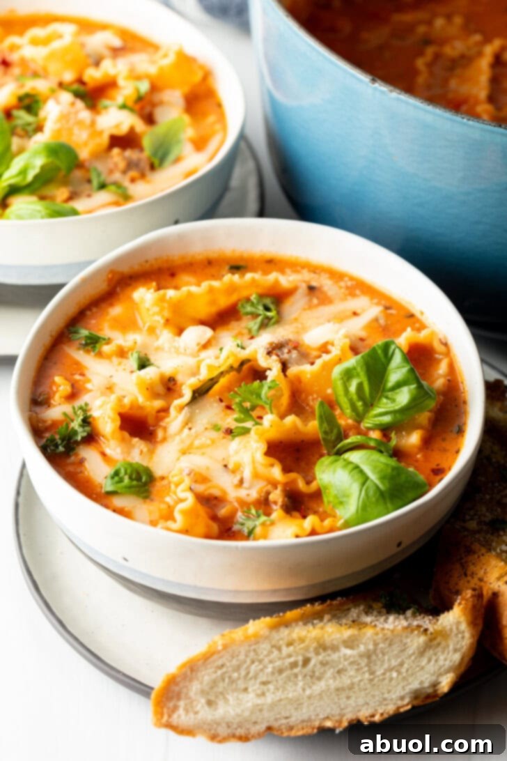 No-Fuss Lasagna Soup 6 White bowl loaded with lasagna soup, garnished with two fresh basil leaves. A slice of baguette is on a white plate under the bowl.