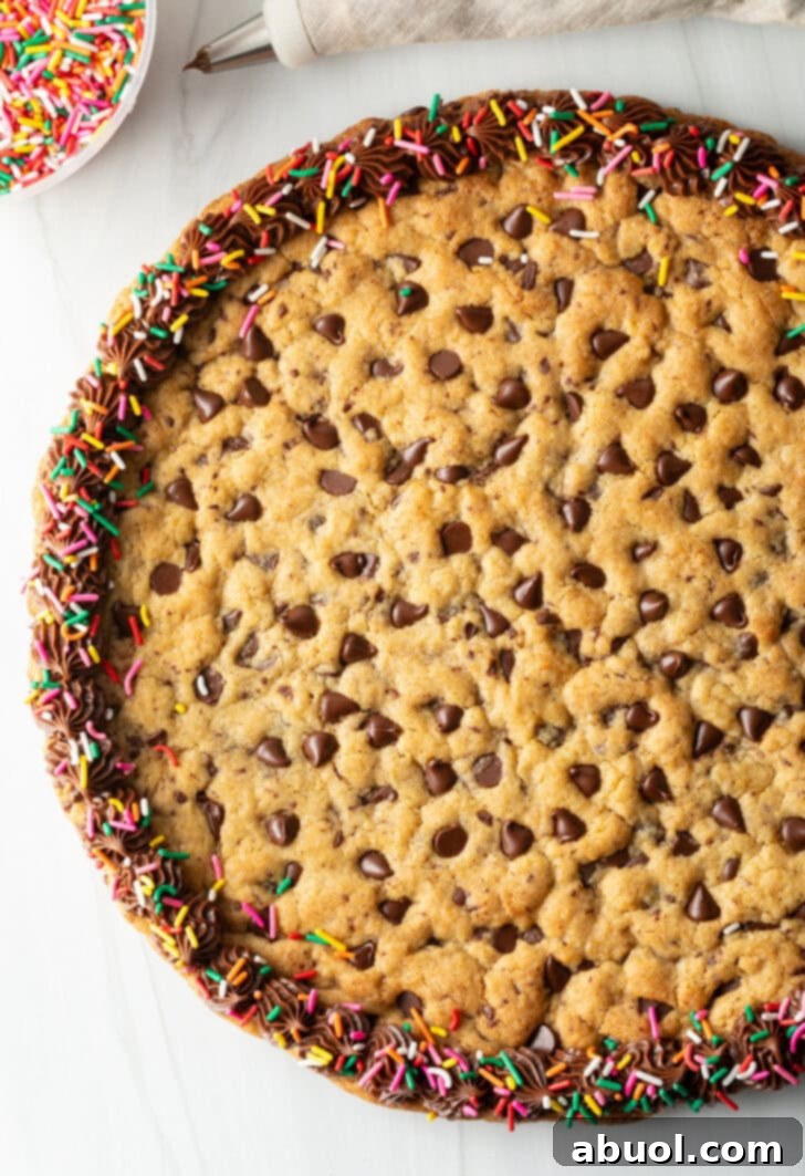 Top down view of a large chocolate chip cookie cake. The outside edge is piped with chocolate frosting and covered with vibrant rainbow sprinkles, making it a festive centerpiece.