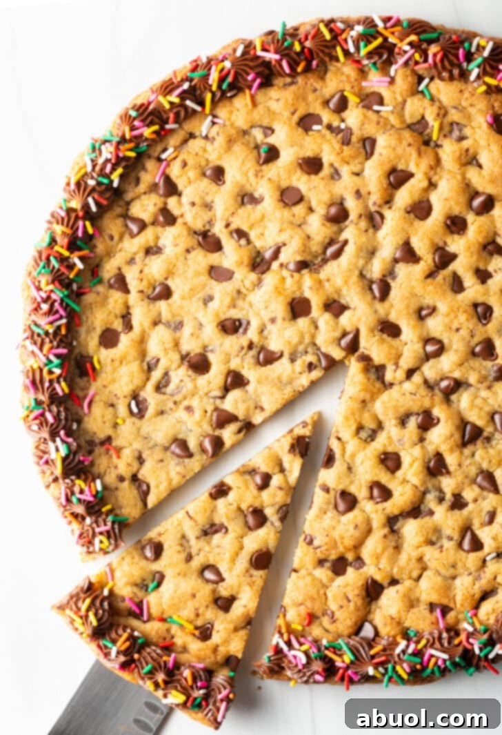 A delicious slice being cut from a large chocolate chip cookie cake. The cake is beautifully decorated with a piped chocolate frosting border and colorful rainbow sprinkles, emphasizing its festive appeal.