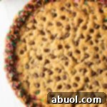Top down view of a large chocolate chip cookie cake. The outside edge is piped with chocolate frosting and covered with rainbow sprinkles.