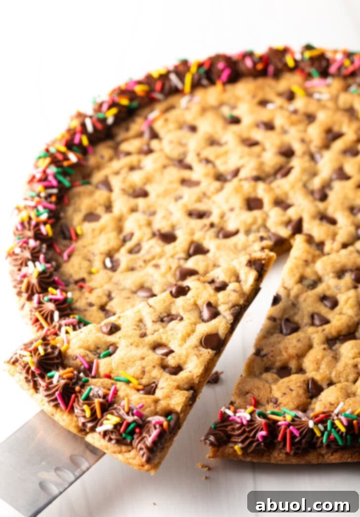 A large chocolate chip cookie cake, exquisitely decorated with a chocolate frosting border and colorful rainbow sprinkles. A cake cutter is poised to take a delicious wedge slice, highlighting its readiness for serving.