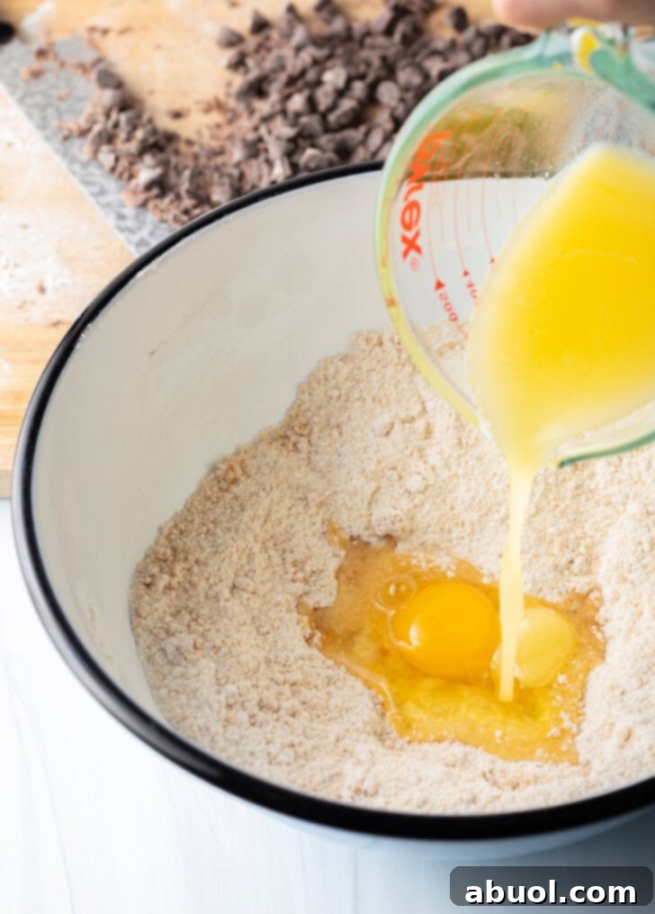 The process of making cookie dough, with melted butter being carefully added to a white mixing bowl containing the dry ingredients. This step ensures proper incorporation for a smooth batter.