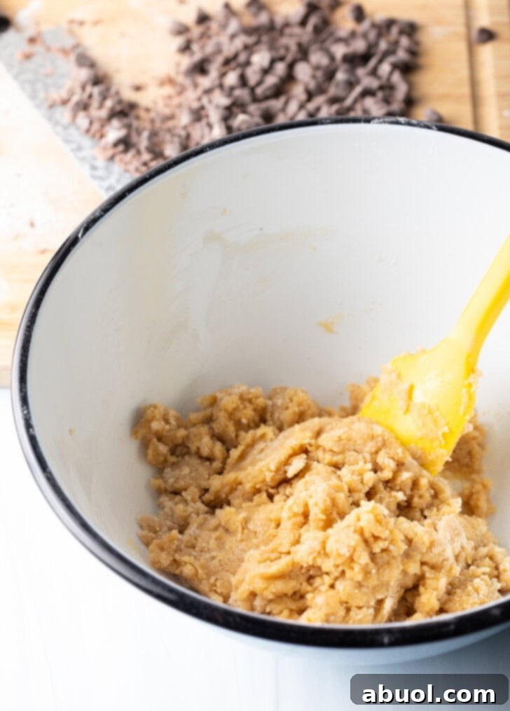 A white mixing bowl holds the creamy cookie dough, being expertly stirred by a yellow spatula, showcasing its smooth consistency before the addition of chocolate chips.