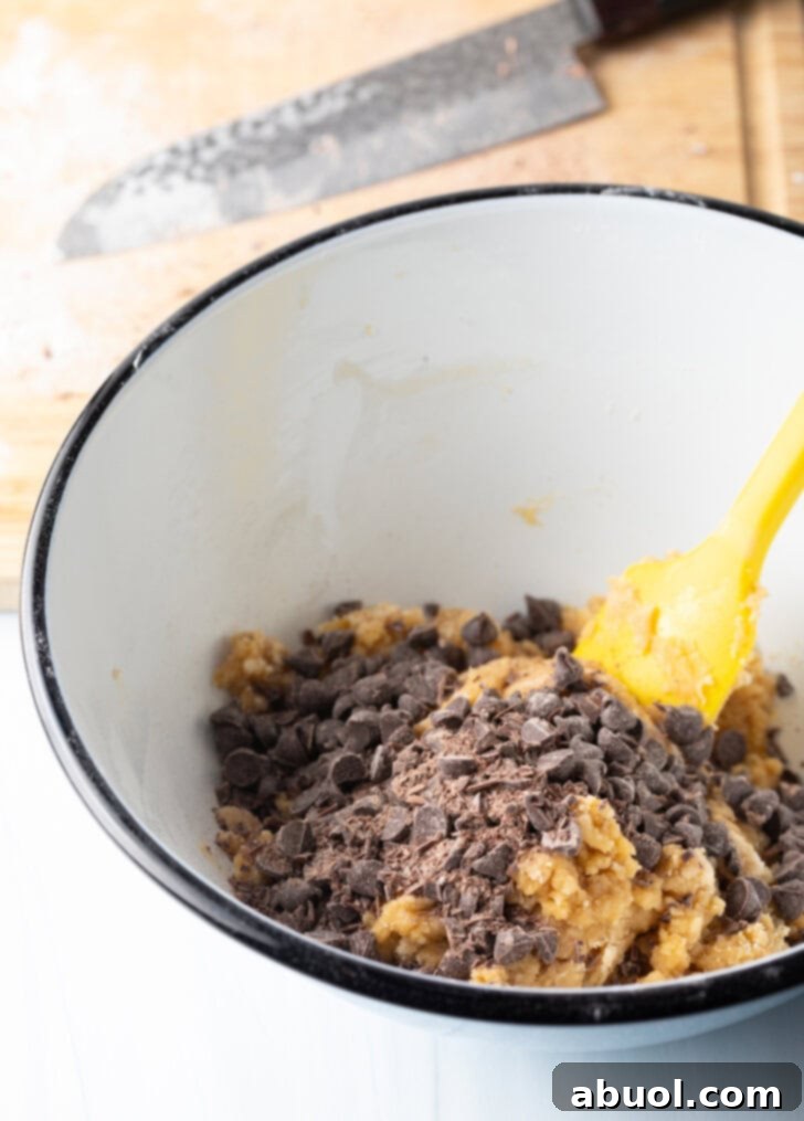 Close-up shot of a white mixing bowl filled with cookie dough and chopped chocolate chips, being mixed with a yellow spatula. This highlights the integration of chocolate into the delicious batter.