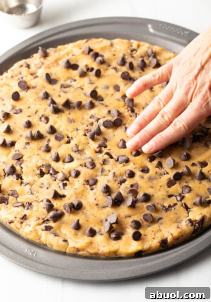 Hands meticulously spreading out the large chocolate chip cookie dough evenly in a pizza pan, creating a smooth, round base for baking.