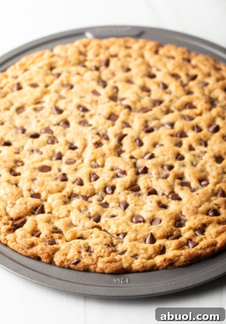 A large, golden-brown chocolate chip cookie cake, freshly baked, sits in its pizza pan, ready to be cooled and decorated.