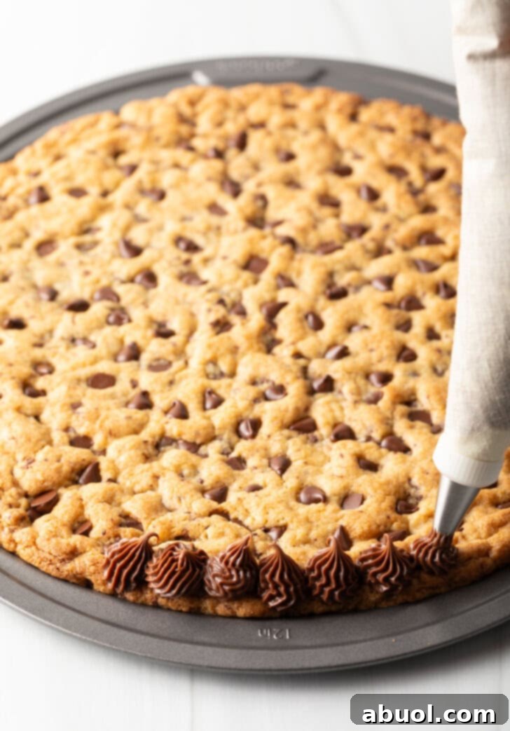 Hands expertly piping a decorative border of rich chocolate frosting along the edge of the large chocolate chip cookie cake, preparing it for festive sprinkles.